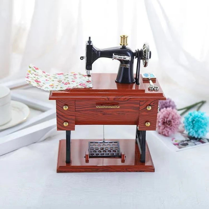 A miniature black sewing machine with a gold detail sits on a wooden box, set against a white background.