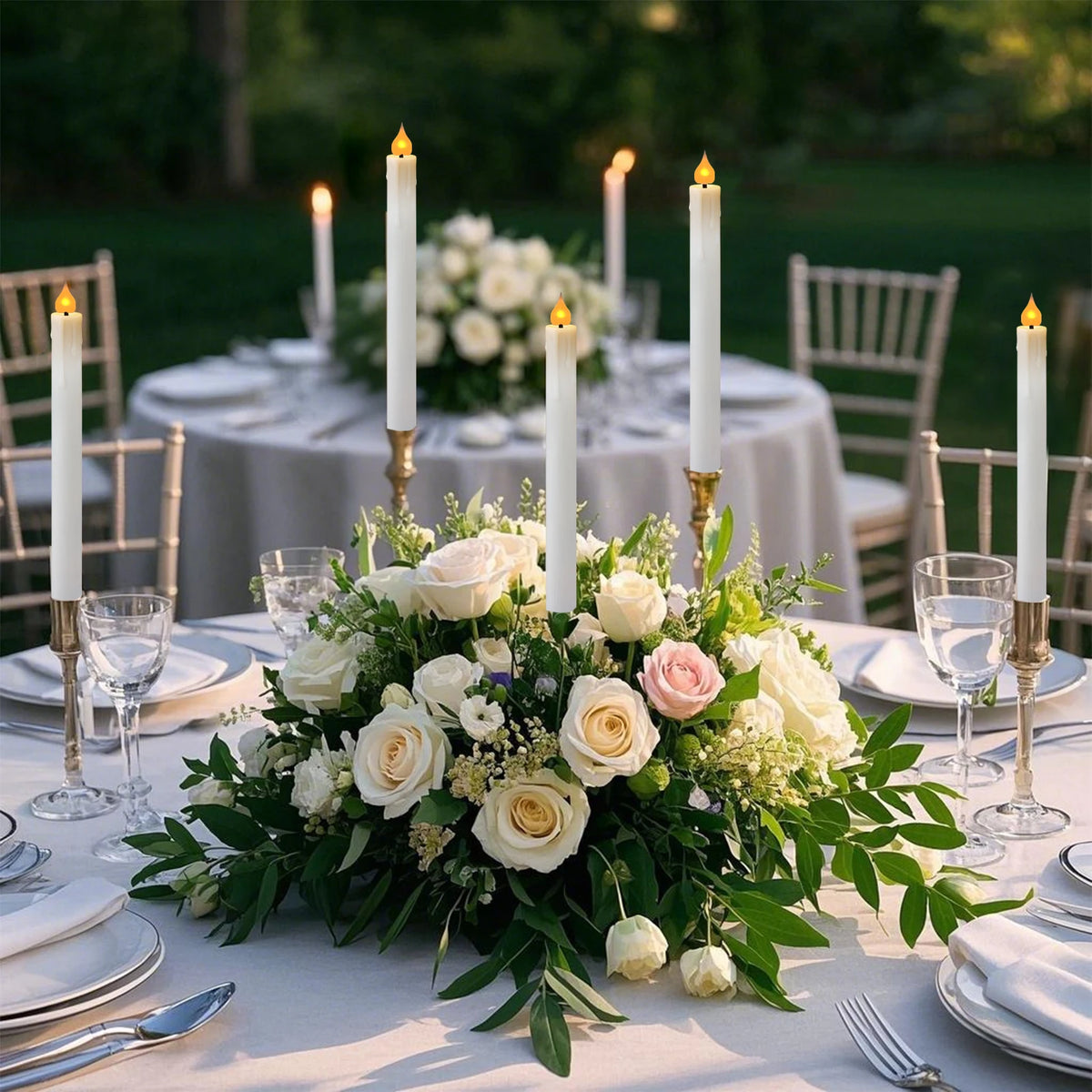 A table setting featuring white taper candles in gold holders surrounded by fresh flowers and greenery.