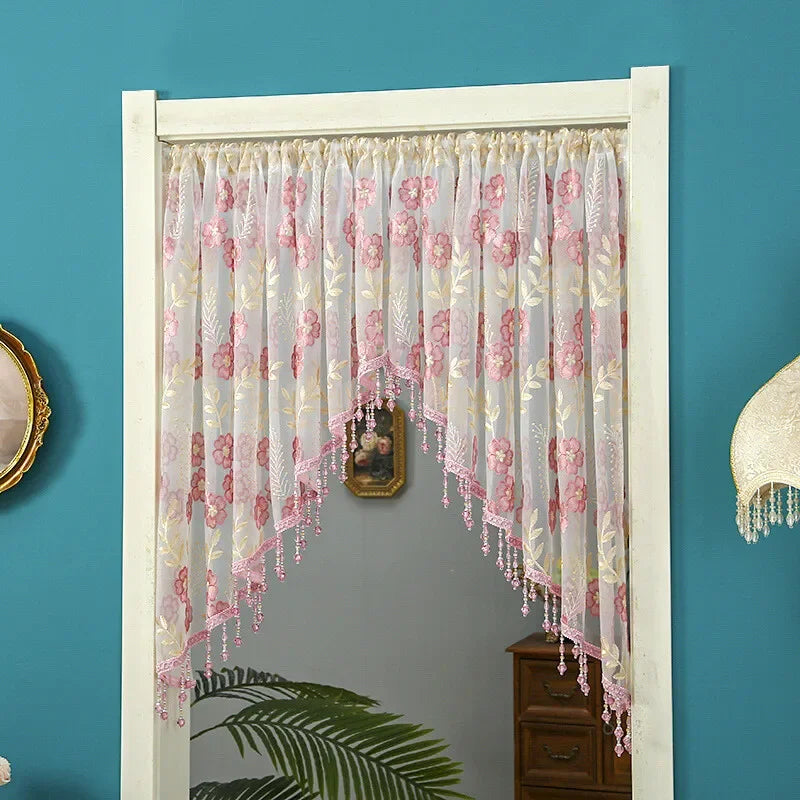 A sheer lace door curtain with pink floral embroidery and tassels hangs in a white frame against a teal wall.