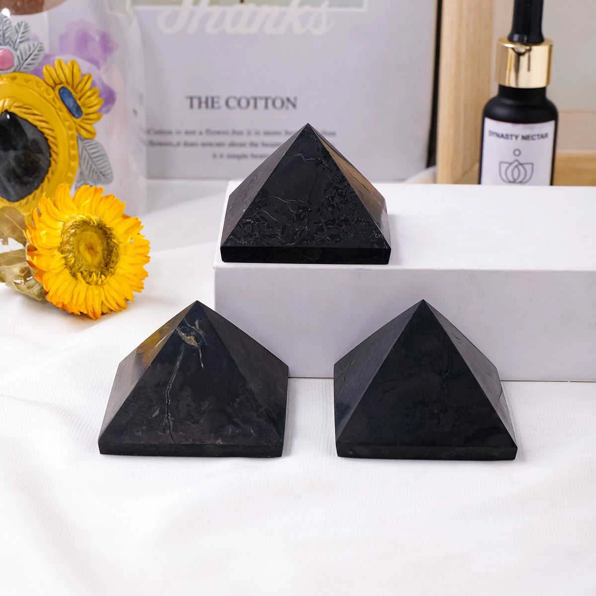 Tres pirámides de cristal de shungita negras exhibidas sobre una superficie blanca junto a una botella de néctar.