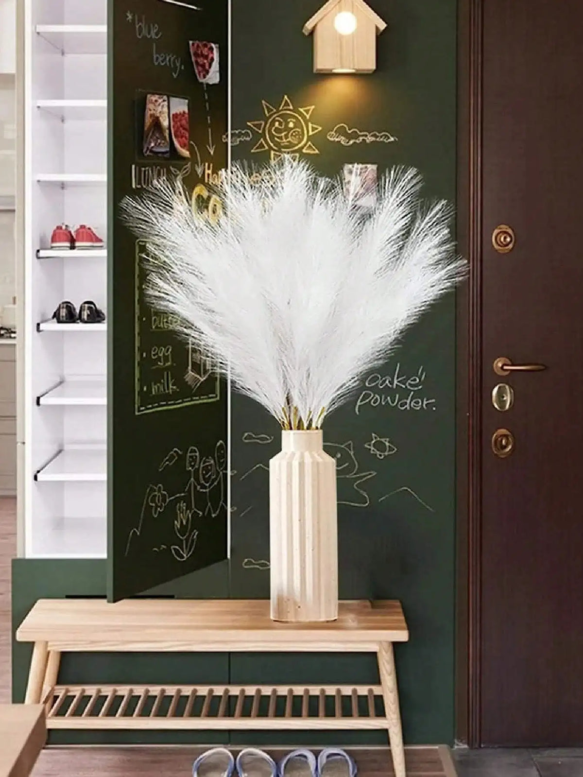 A vase holds a bouquet of white pampas grass stems displayed on a green cabinet door.