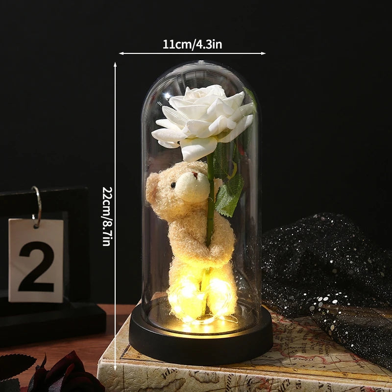 A glass dome display featuring a white rose and a small teddy bear, illuminated from within.