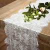 Un camino de mesa de encaje floral vintage con hojas verdes y flores blancas sobre una mesa de madera.