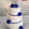 A three-tiered white cake decorated with blue roses and silver accents, set against a sparkling background.