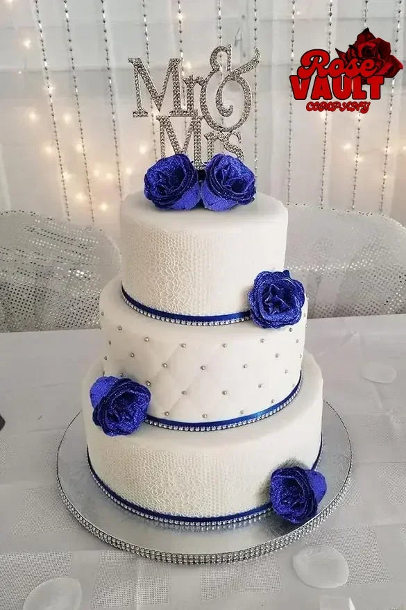 A three-tiered white cake decorated with blue roses and silver accents, set against a sparkling background.