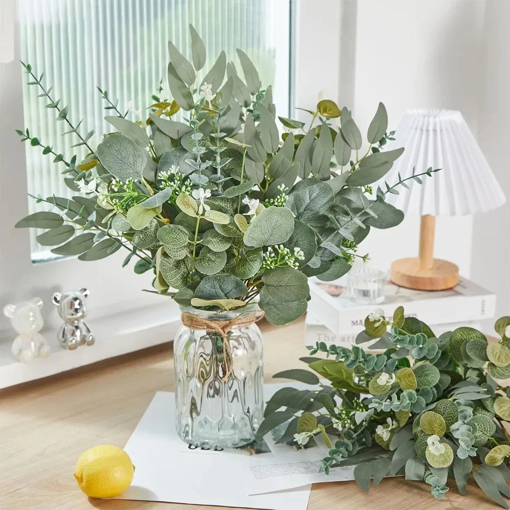 A bouquet of artificial eucalyptus leaves in various shades of green displayed in a glass jar on a wooden surface.