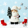 Figura de Papá Noel con barba y sombrero blanco junto a un árbol verde rodeado de copos de nieve en una superficie nevada.