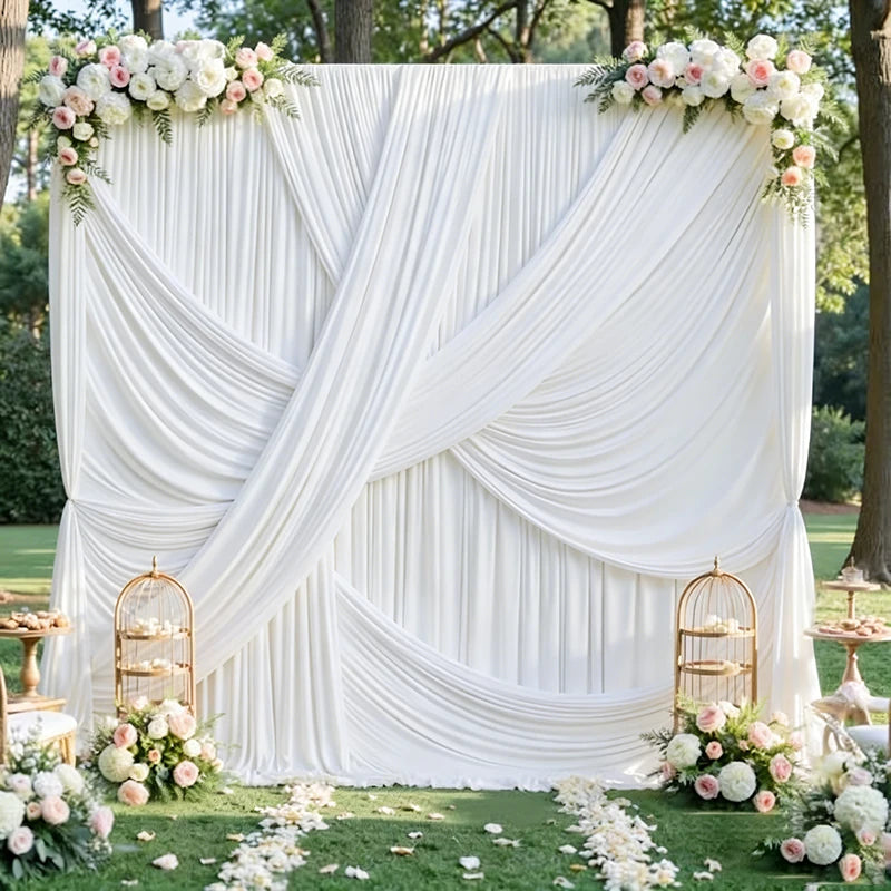 A white fabric backdrop curtain draped in soft folds and adorned with floral greenery on a grassy outdoor setting.