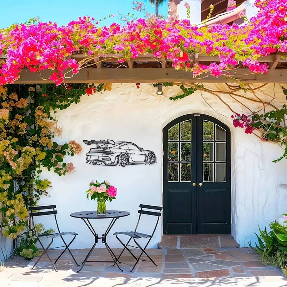 Black sports car metal wall art displayed on a white stucco wall with pink and green foliage.