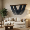 A bohemian macrame wall tapestry hangs on a beige wall above a light-colored sofa and plant.