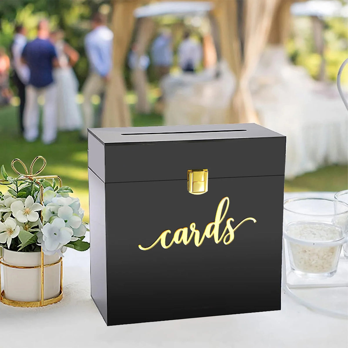 A black acrylic gift box with gold lettering labeled cards sits on a white surface in an outdoor event setting.