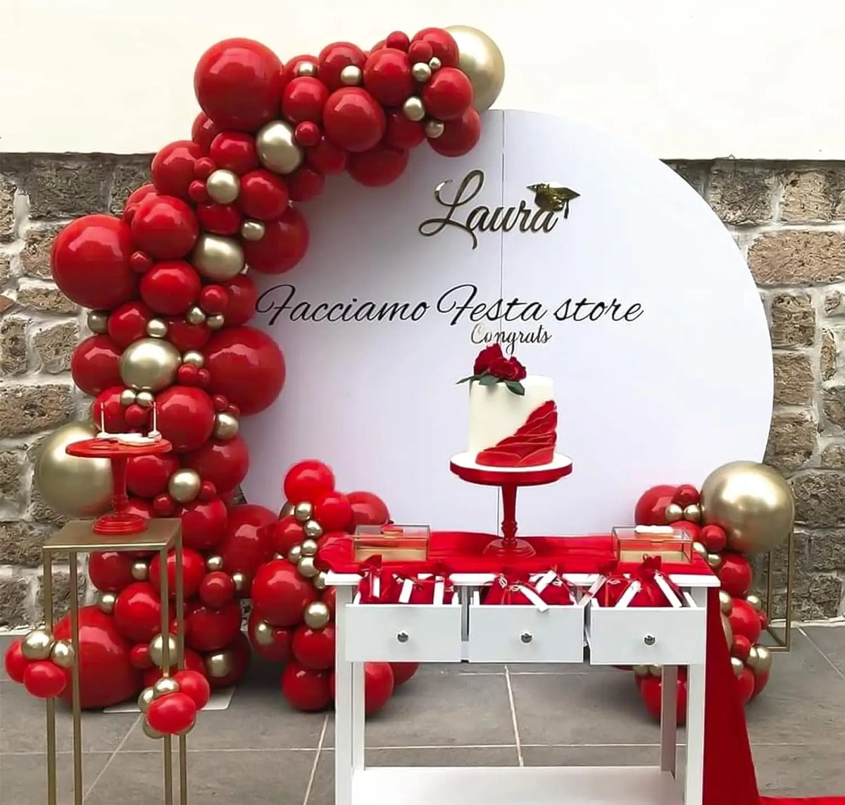 A red and gold balloon arch decoration featuring a white cake display against a stone wall.