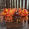 Lebendige Anordnung künstlicher Herbstblumen in Orange-, Rot- und Gelbtönen in einer Holzbox.