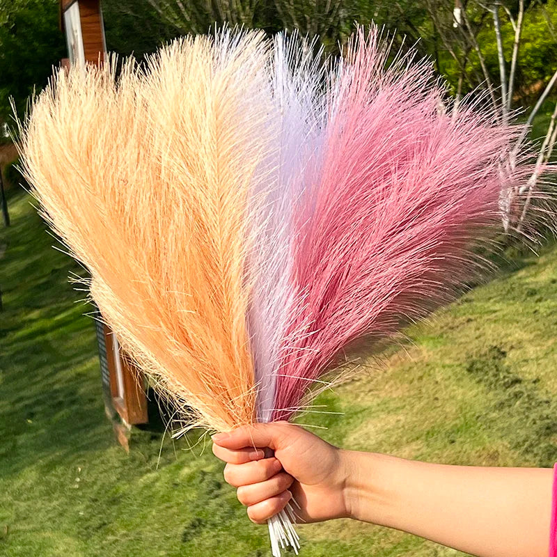A hand holds a bouquet of artificial reed flowers in shades of orange, pink, and purple.