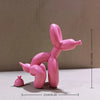 A pink balloon dog sculpture in a squatting pose, displayed on a white surface.