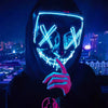 A person wearing a glowing blue LED mask with an 'X' design, holding a finger to the mask in a dark city setting.