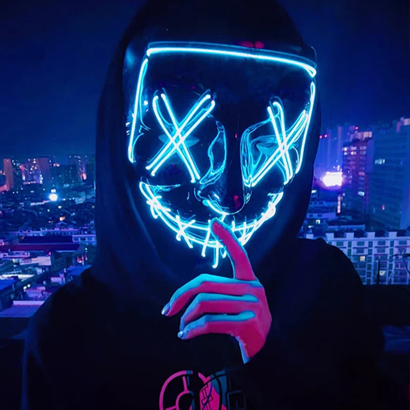 A person wearing a glowing blue LED mask with an 'X' design, holding a finger to the mask in a dark city setting.
