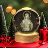 A glass crystal ball night light featuring a figure of Jesus, set on a wooden base and surrounded by green pine trees.