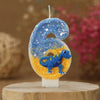 A birthday candle shaped like the number six decorated with blue glitter and a blue dinosaur silhouette on an orange backgrou