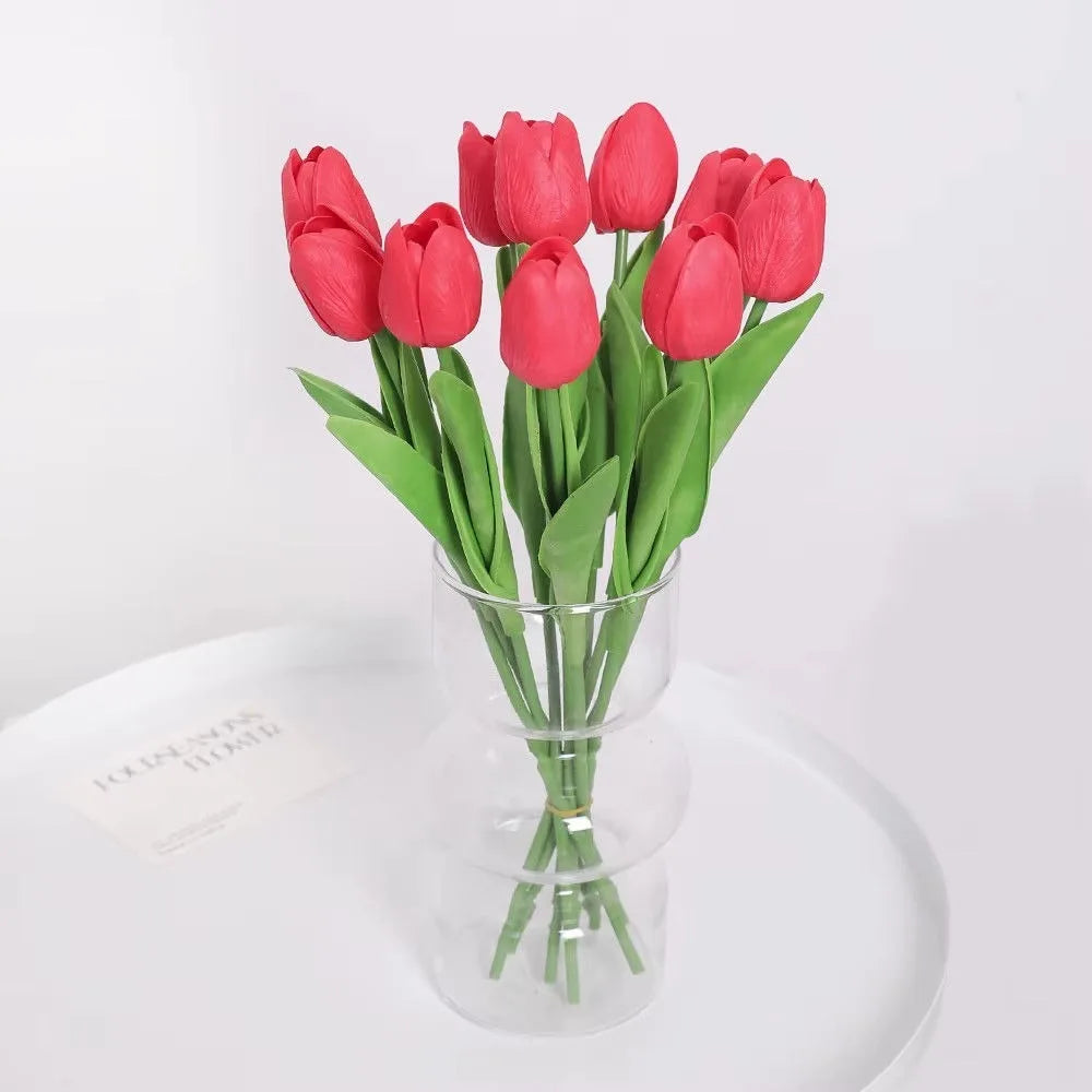 A bouquet of bright pink artificial tulip flowers with green stems in a clear glass vase.