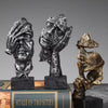 A set of three resin figurines depicting faces covered in hands, displayed on a book.