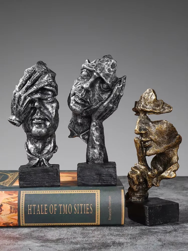 A set of three resin figurines depicting faces covered in hands, displayed on a book.