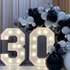 A large light-up marquee number sign displaying the number 30, surrounded by a balloon garland of black and white spheres.