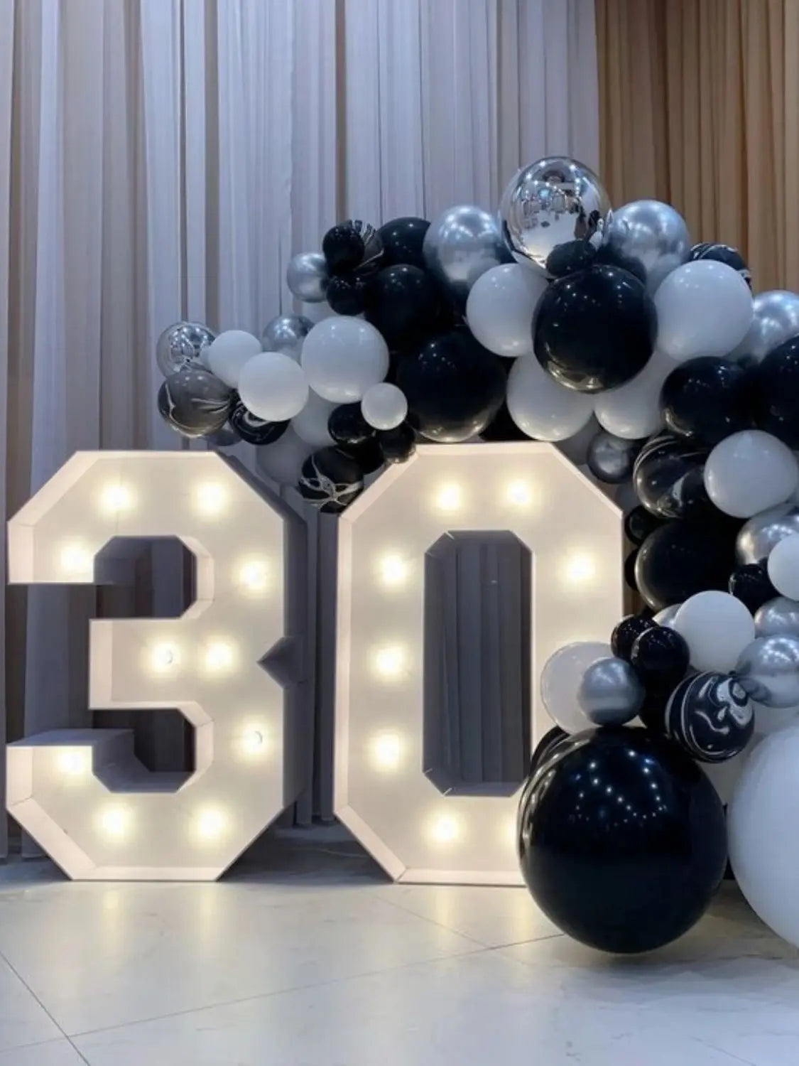 A large light-up marquee number sign displaying the number 30, surrounded by a balloon garland of black and white spheres.