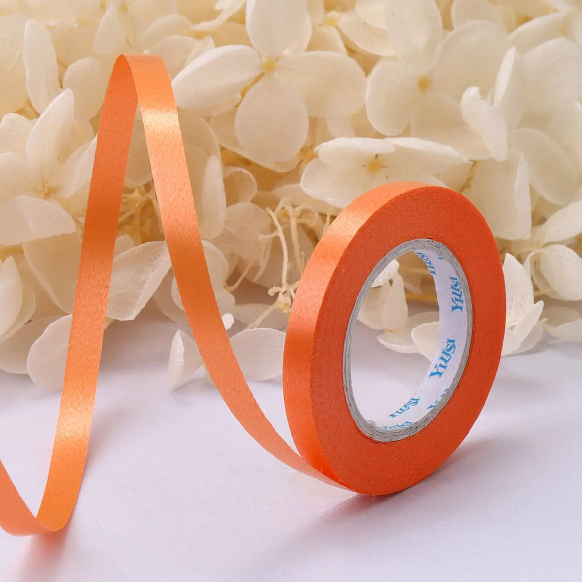 Orange satin balloon ribbon roll unwinding against a background of white flowers.