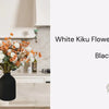 A large matte black decorative vase filled with white and orange wax flowers on a light-colored kitchen counter.