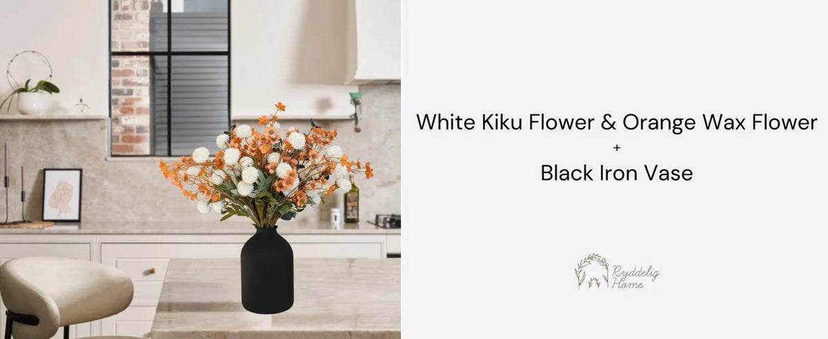 A large matte black decorative vase filled with white and orange wax flowers on a light-colored kitchen counter.
