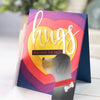 A greeting card with the word hugs in gold script on a colorful background and a silhouette of a dog's head.