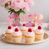 A display of pink and glitter crown and bow cupcake toppers arranged on a gold platter with pink cupcakes and flowers in the 
