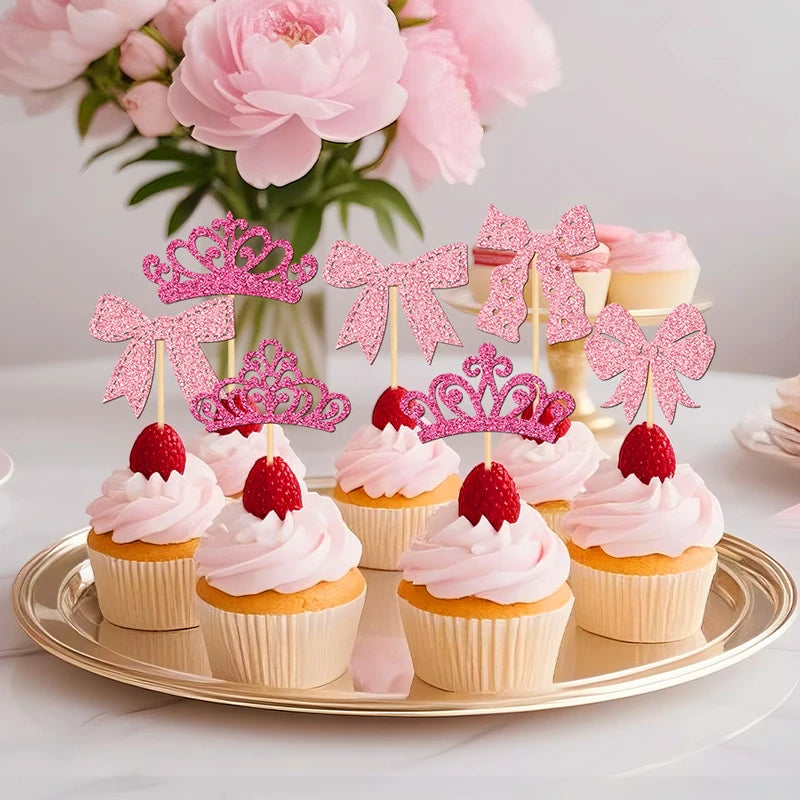A display of pink and glitter crown and bow cupcake toppers arranged on a gold platter with pink cupcakes and flowers in the 