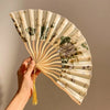 A hand holding a light beige folding fan with a leopard print design and black handle.