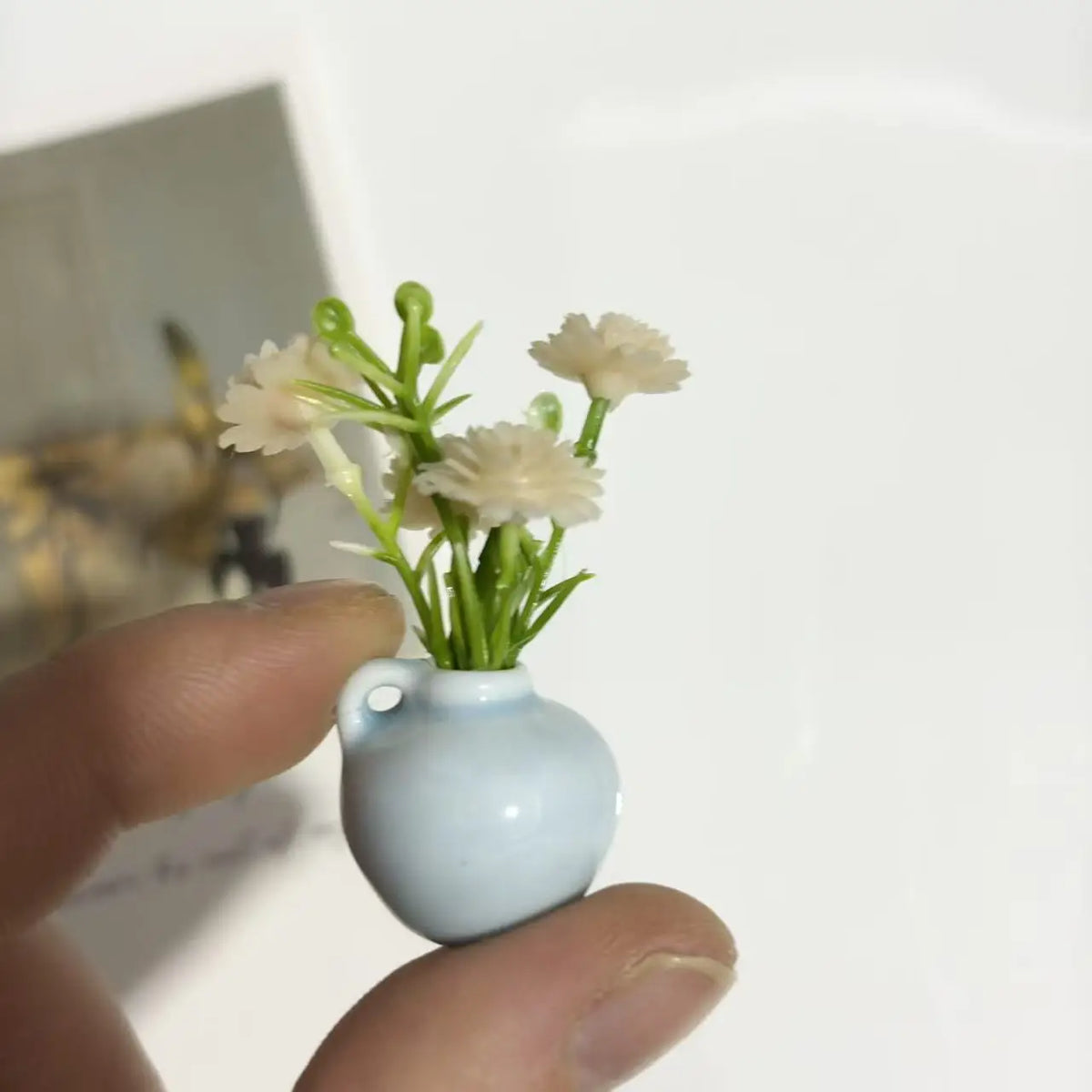 A hand holds a small, light blue ceramic vase with artificial white flowers.