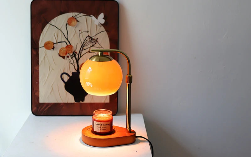 A golden aromatherapy wax melting lamp with a warm glow sits on a white surface next to a framed botanical print.