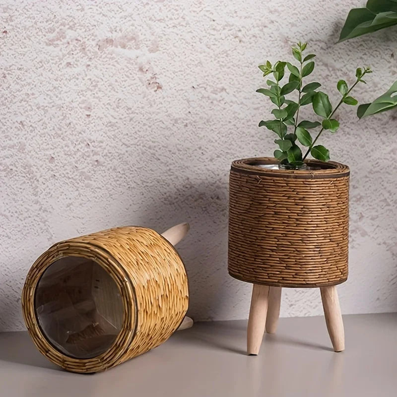 A rustic rattan planter with a wooden lid and legs holding a green plant against a textured wall.