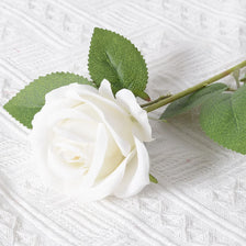 A single white artificial rose with green leaves resting on a white textured fabric.