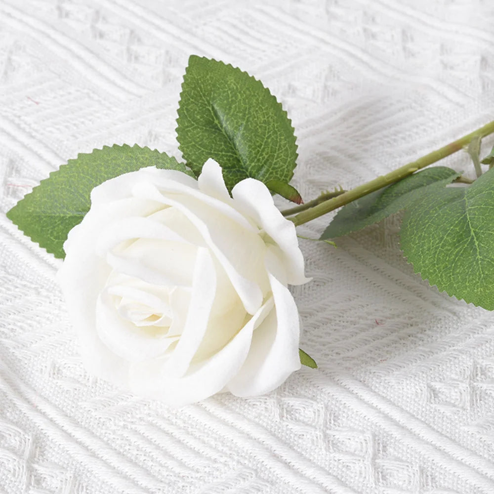 A single white artificial rose with green leaves resting on a white textured fabric.