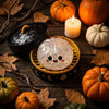 A glowing ghost dumpling toy sits in a dark bowl surrounded by autumn leaves and pumpkins.