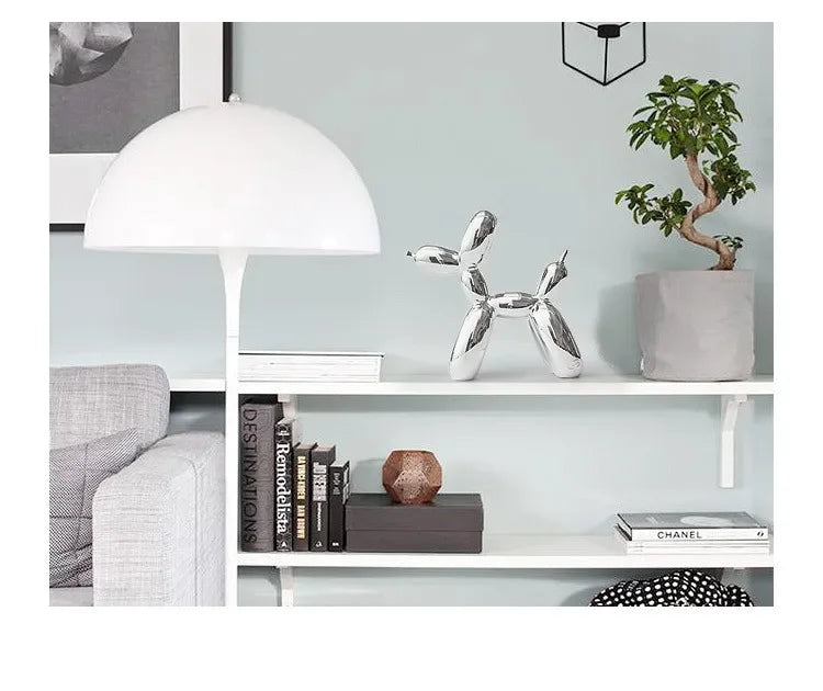A silver resin balloon dog sculpture sits on a white shelf next to a potted green tree and a white lampshade.