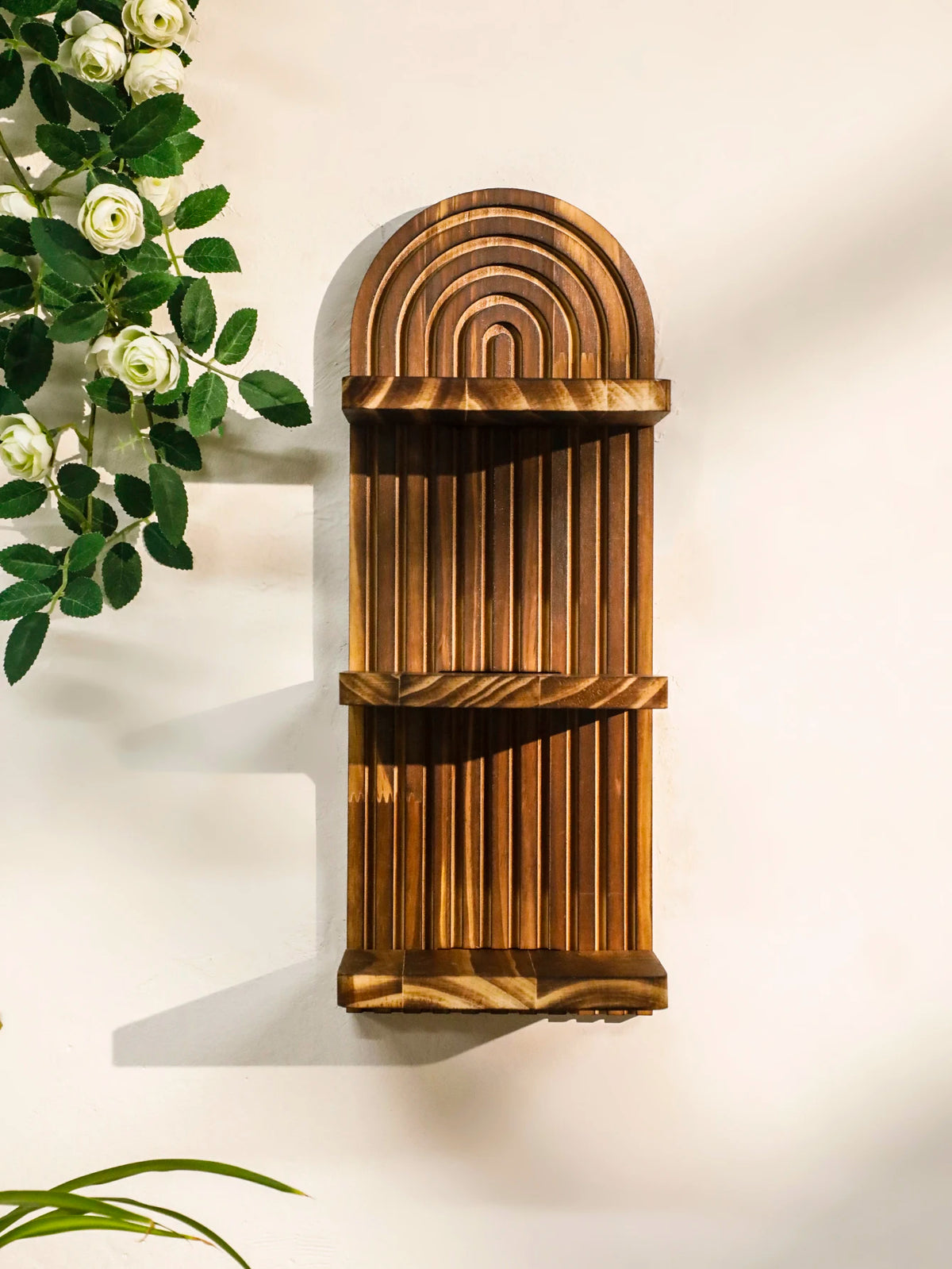 A vertical wooden wall shelf with a curved, ribbed design mounted on a white wall next to green foliage.