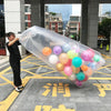 A giant clear plastic bag filled with colorful balloons is being held by a person on a paved outdoor area.