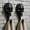 A set of two black gothic skull wall hooks mounted on a gray brick wall, with two beige towels hanging below.