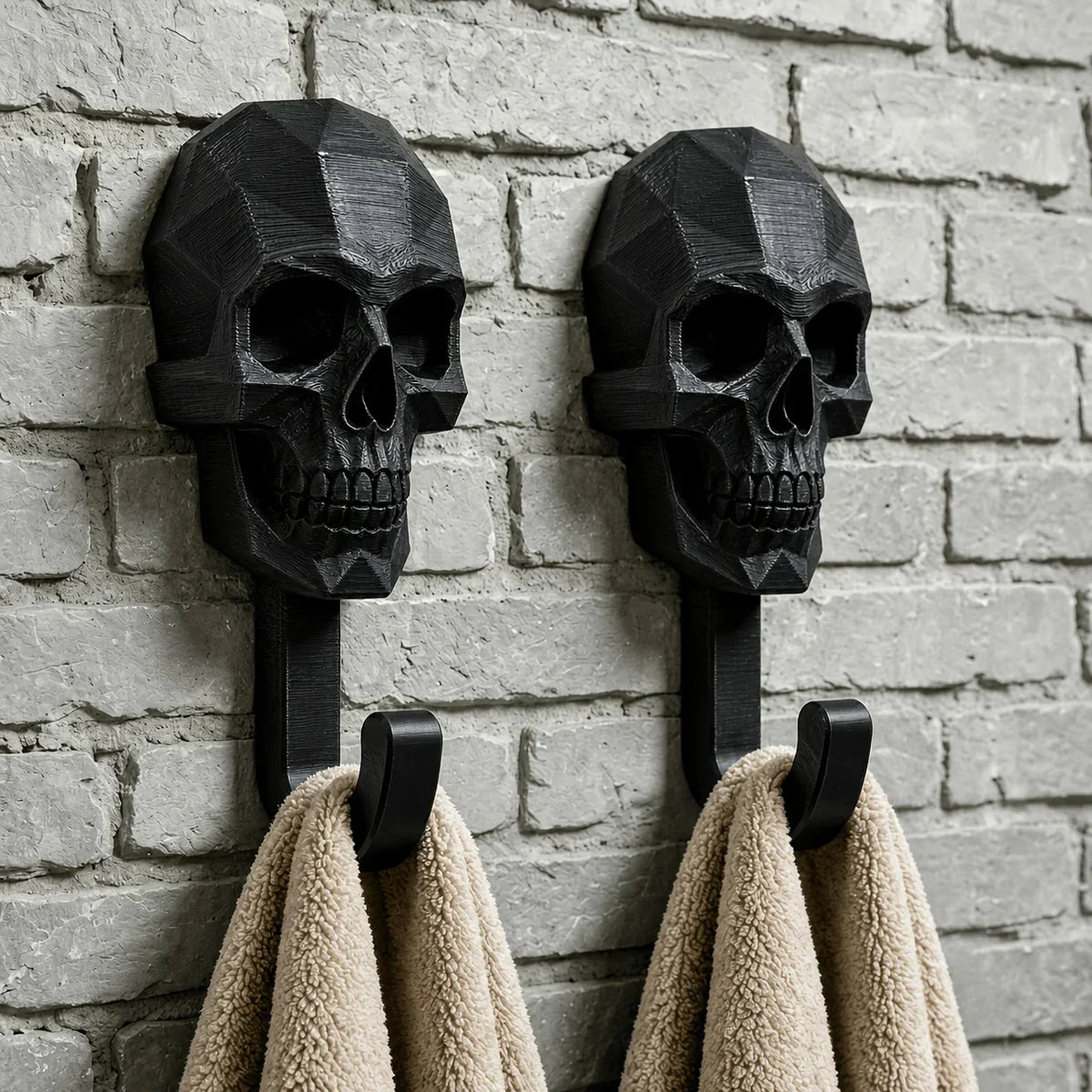 A set of two black gothic skull wall hooks mounted on a gray brick wall, with two beige towels hanging below.