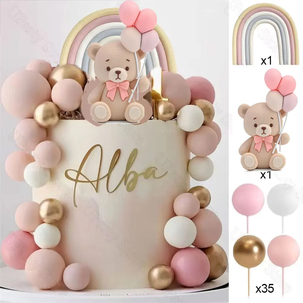 A cake decorated with pink and gold ball decorations and a teddy bear topper, set with rainbow arch.