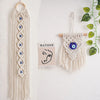 Macrame wall hanging featuring a long fringe and an evil eye pendant on a wooden hanger.