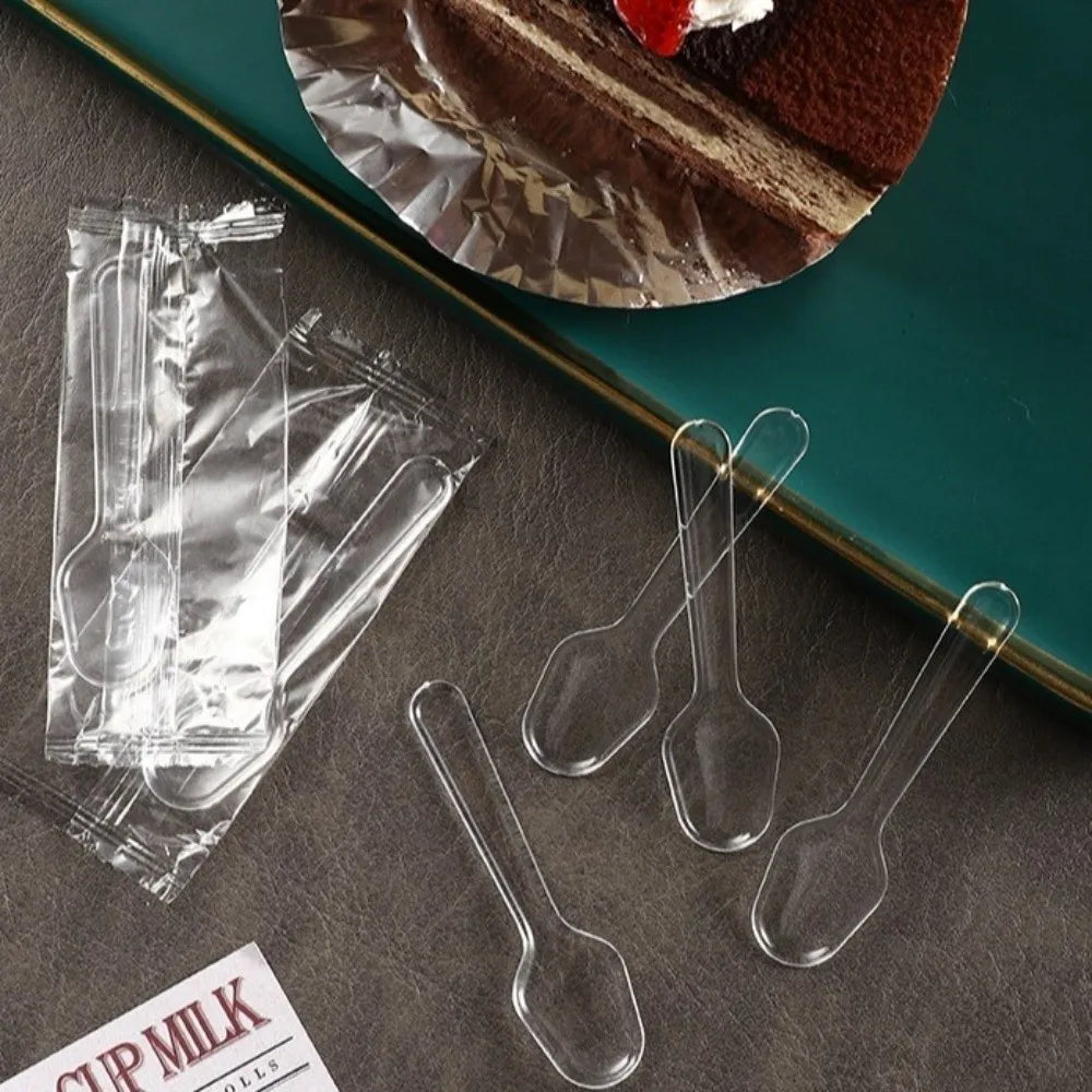 Clear plastic disposable spoons arranged on a dark surface next to a cake and foil.