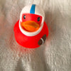 A bright red rubber duck bath toy with an orange beak and blue stripe sitting on a silver textured surface.